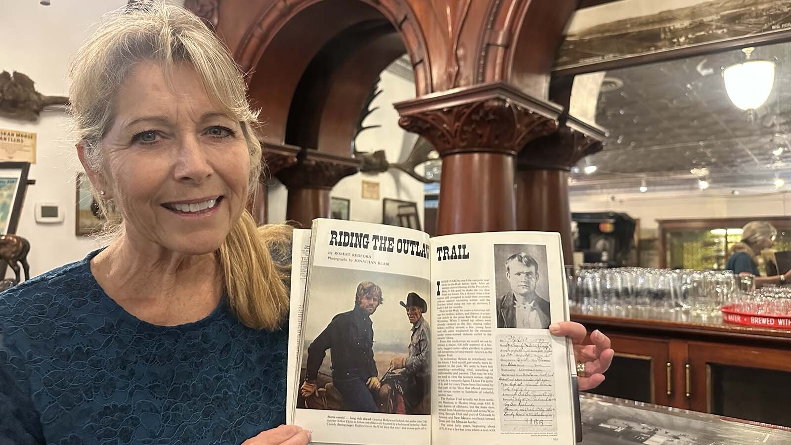 Hot Springs County Museum employee Terri Geissinger poses at the bar that Robert Redford attempted to buy in 1993. She holds the 1976 National Geographic magazine that he wrote the cover article for about his trip along the Outlaw Trail that took him through Wyoming.