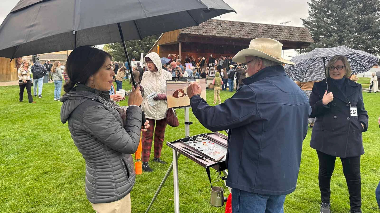 Katelyn Parker, executive director of the Buffalo Bill Art Show and Sale, uses an umbrella to keep artist Steve Devenyns and his oil painting dry during Saturday morning's Quick Draw. 