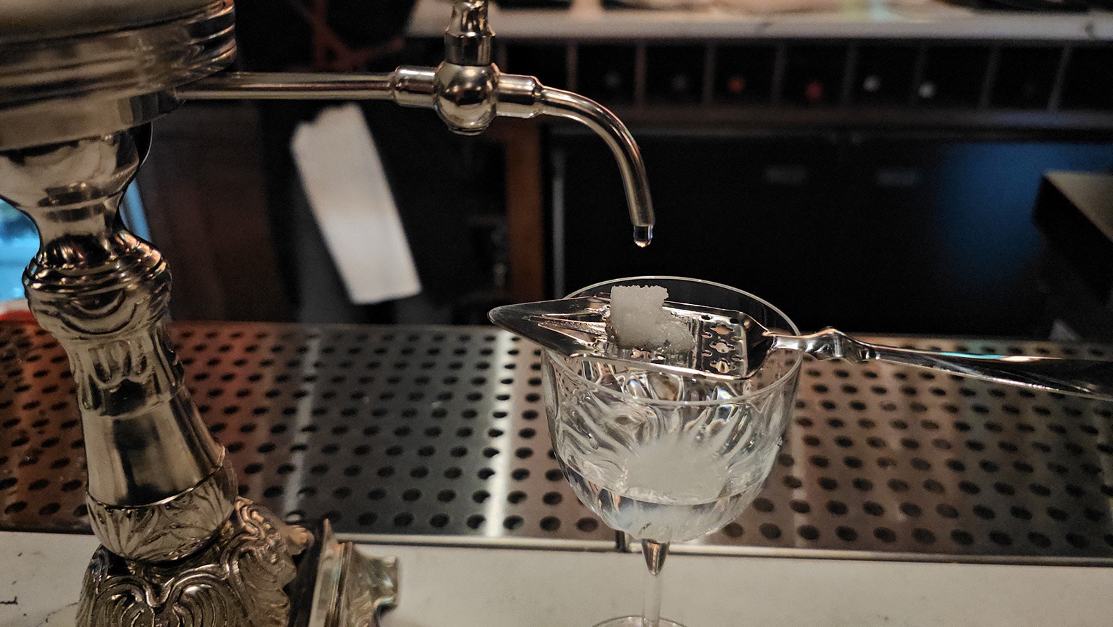 A slotted spoon holds a sugar cube above a shot of absinthe, while water slowly drips down, dissolving the sugar and diluting the alcohol.