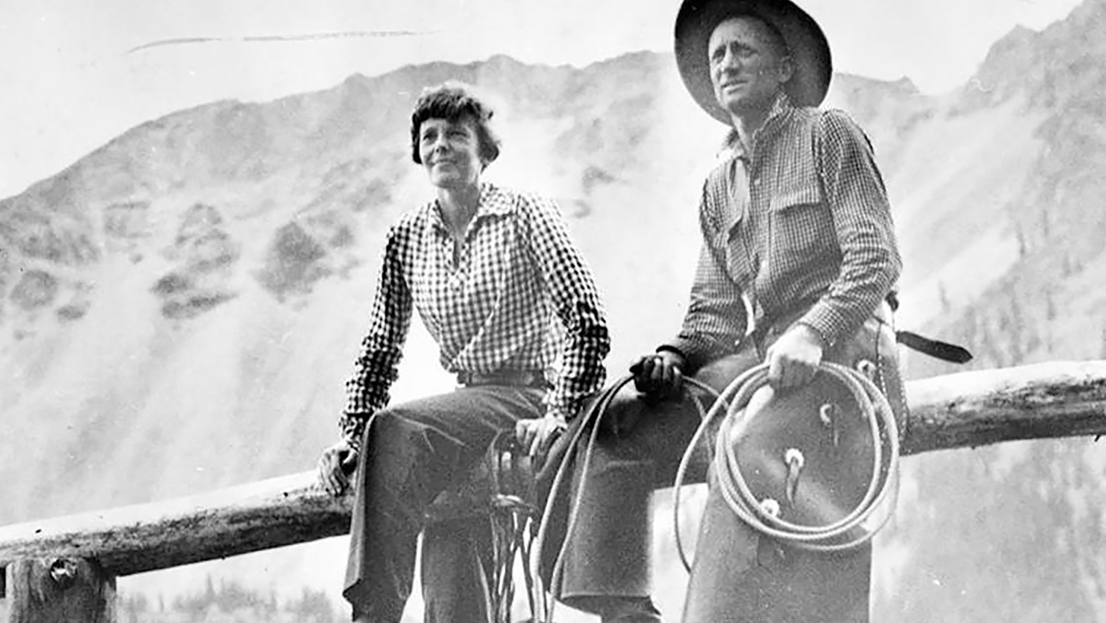 Amelia Earhart and Carl Dunrud sit on the top rail of a fence at Double D Ranch on the upper Wood River in Wyoming with the Absaroka Mountains in the background.