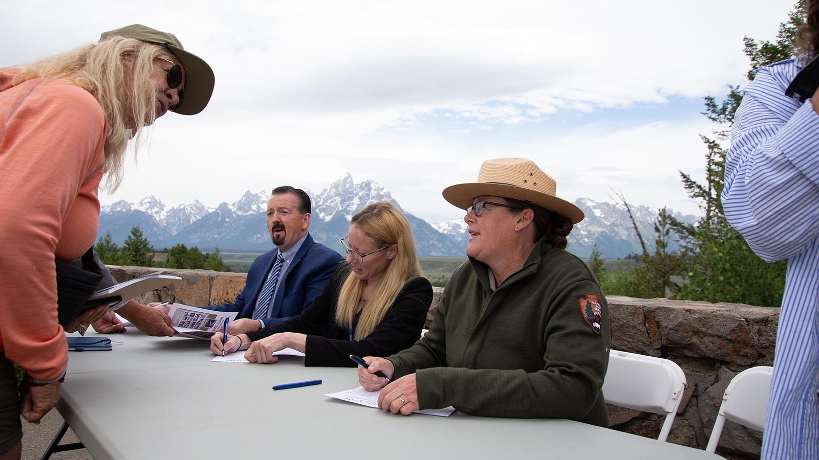 U.S. Postal Service District Manager Douglas Smith, from left, Moose Postmaster Aleicia Dickson and Grand Teton National Park Acting Deputy Superintendent Park Robin Martin sign programs for attendees at the reveal of the Ansel Adams stamp series with the Grand Tetons in the background.