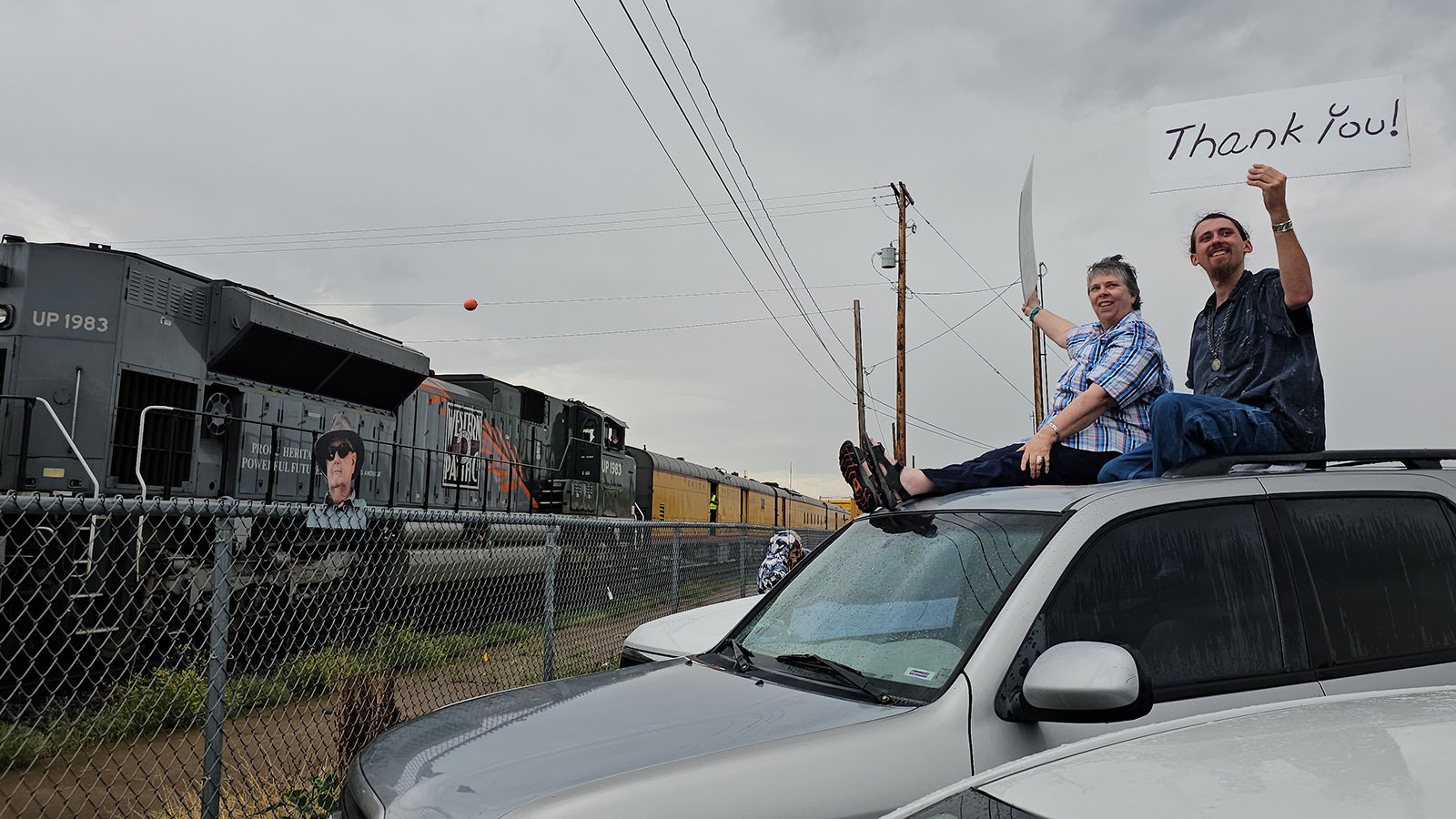 Sue Connelly and Cimarron Jeremiah hold up "thank you" signs as Big Boy 4014 rolls into Cheyenne, with a life-size photographic cutout of their friend Kirk Weilepp, who was a big train chaser until his death earlier this year from pancreatic cancer.