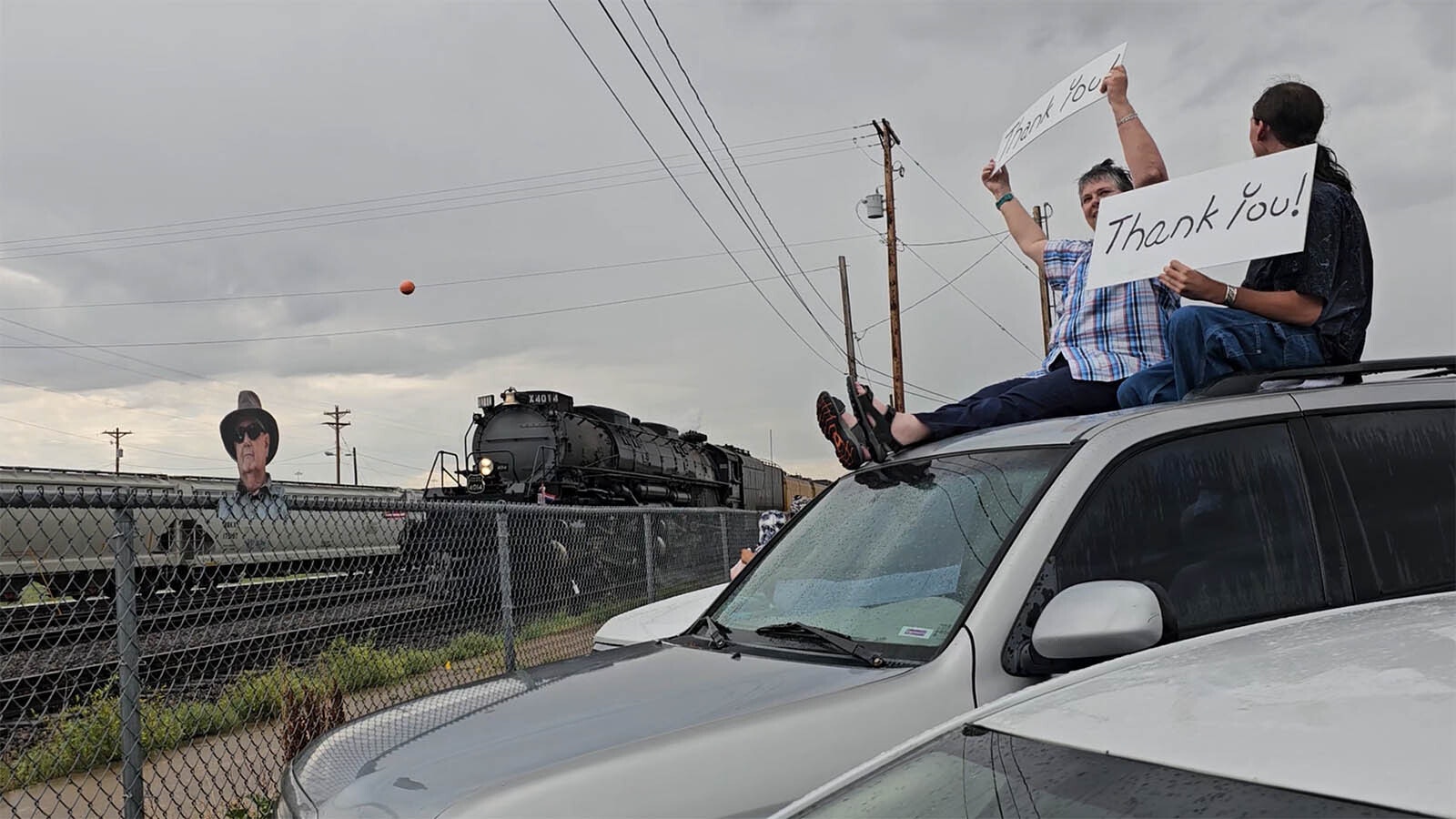 Sue Connelly and Cimarron Jeremiah hold up "thank you" signs as Big Boy 4014 rolls into Cheyenne, with a life-size photographic cutout of their friend Kirk Weilepp, who was a big train chaser until his death earlier this year from pancreatic cancer.