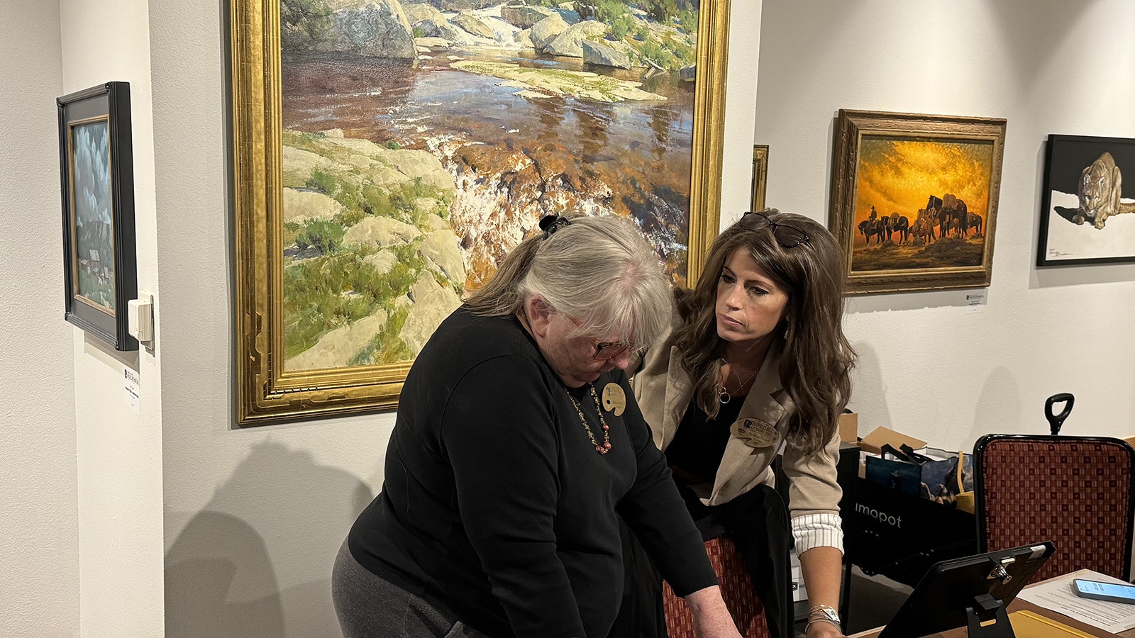 Katelyn Parker, director of the Buffalo Bill Art Show and Sale, shows a concierge how to take payments for paintings in the gallery of the 43rd Buffalo Bill Art Show and Sale. Behind them is "The Last Half-Mile," an oil painting by Kimball Geisler.