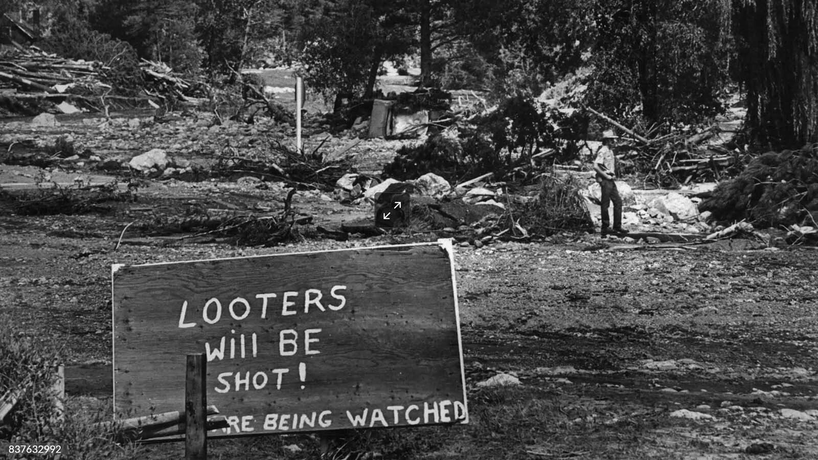 Technician David Smelter with the Colorado Division of Wildlife stands guard against looters in flood-devastated town of Drake in Big Thompson Canyon on Aug. 5, 1976. The sign in foreground expresses feelings of local residents, but Smelter said looting hasn't been problem.