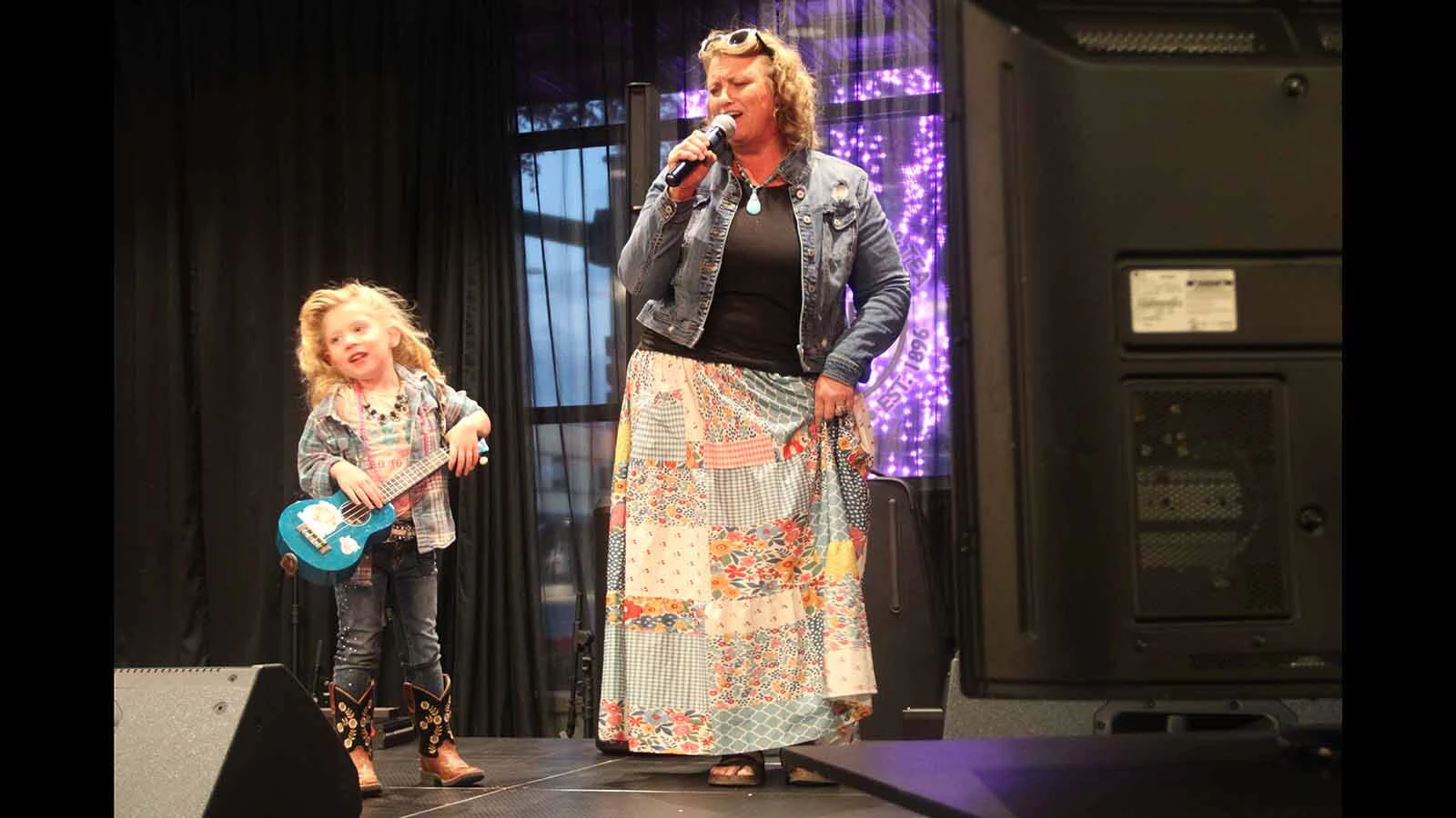 A karaoke singer shares the stage with a young participant holding a blue ukulele during Casper’s Dolly Day celebration Friday.