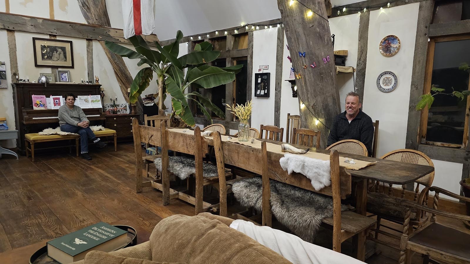 Cyning Meadowcroft sits at his dining hall table, whose benches are made from wood that came from a cottage dating back to the 1000s. His wife, Christine, sits to the left, on a piano bench.