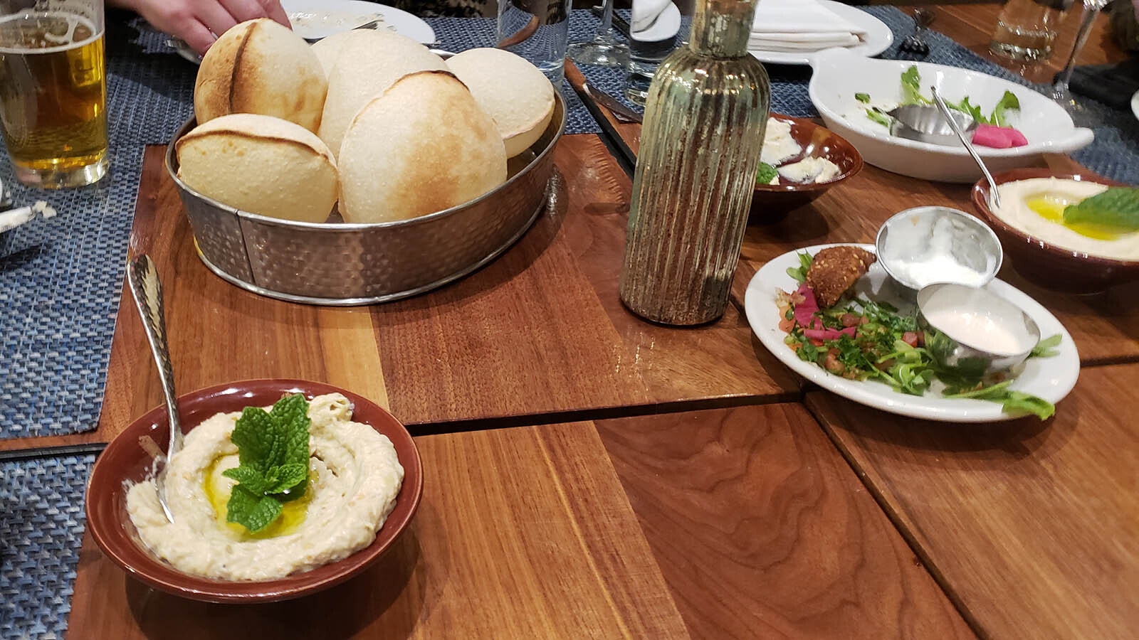 Hummus comes drizzled with olive oil and garnished with mint In the background, a tin basket full of toasted balloon bread or puff pitas.