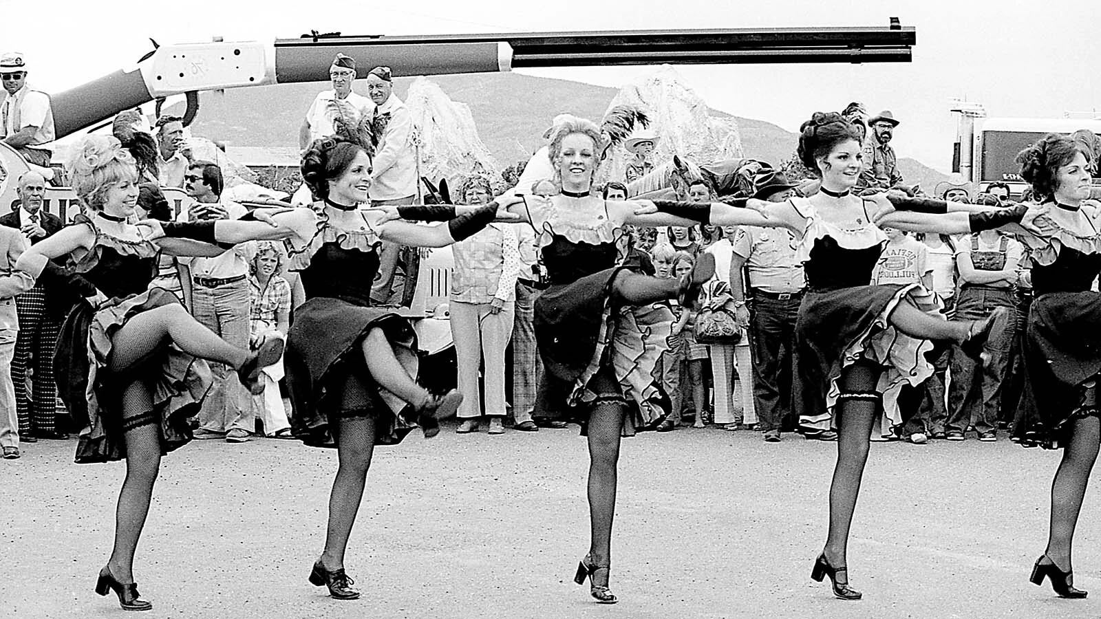 Katy Brown's Buffalo Bill Dollies can-can girls dance with the giant Winchester rifle behind them in Cody, Wyoming, during the rifle's debut in 1976. The scene is the Cody airport terminal parking lot while waiting for John Wayne's ceremonial arrival.