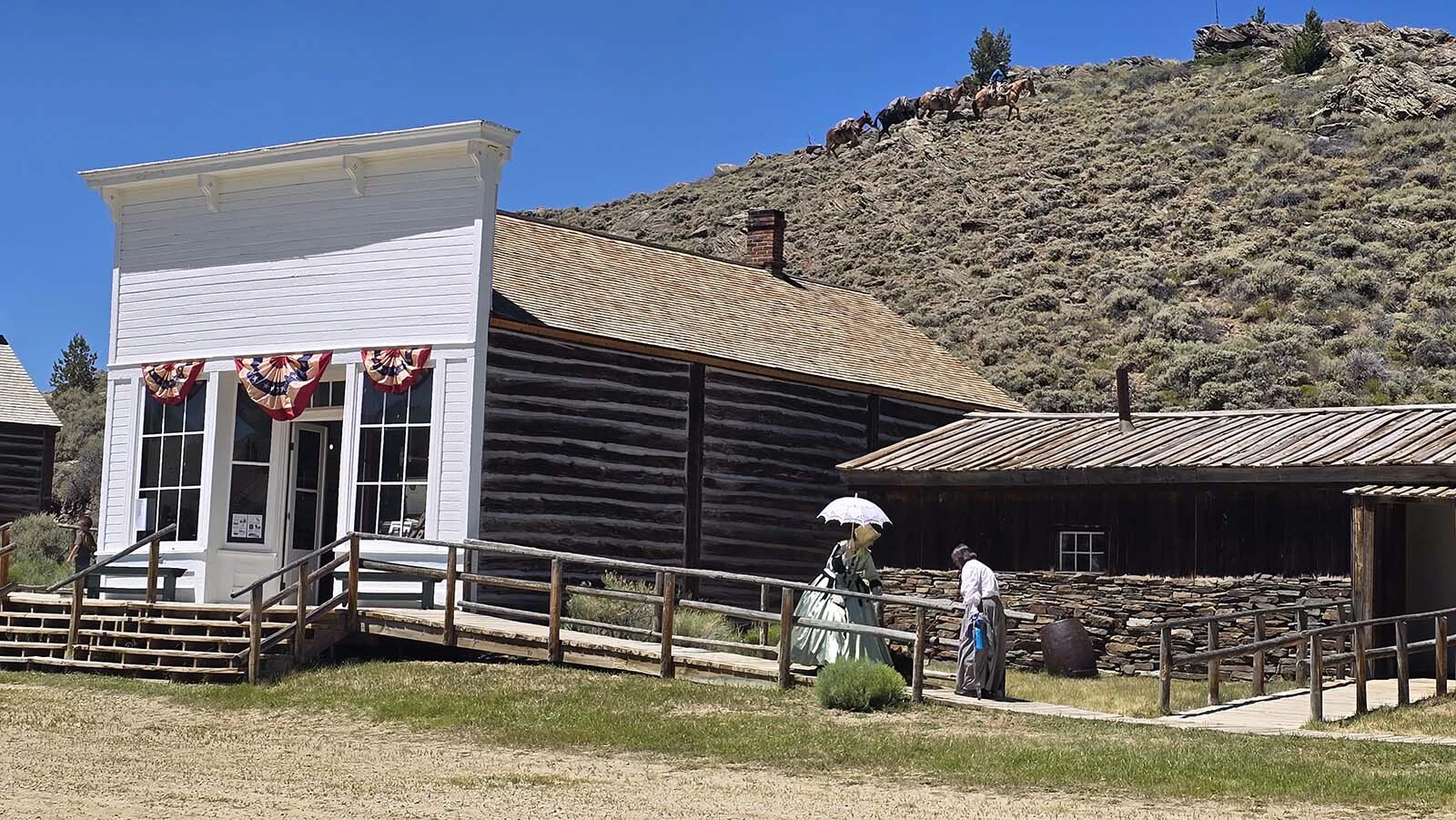 A pack mule team climbs the hill behind South Pass City while a pair of visitors dressed in period attire walk to the Sherlock Store.