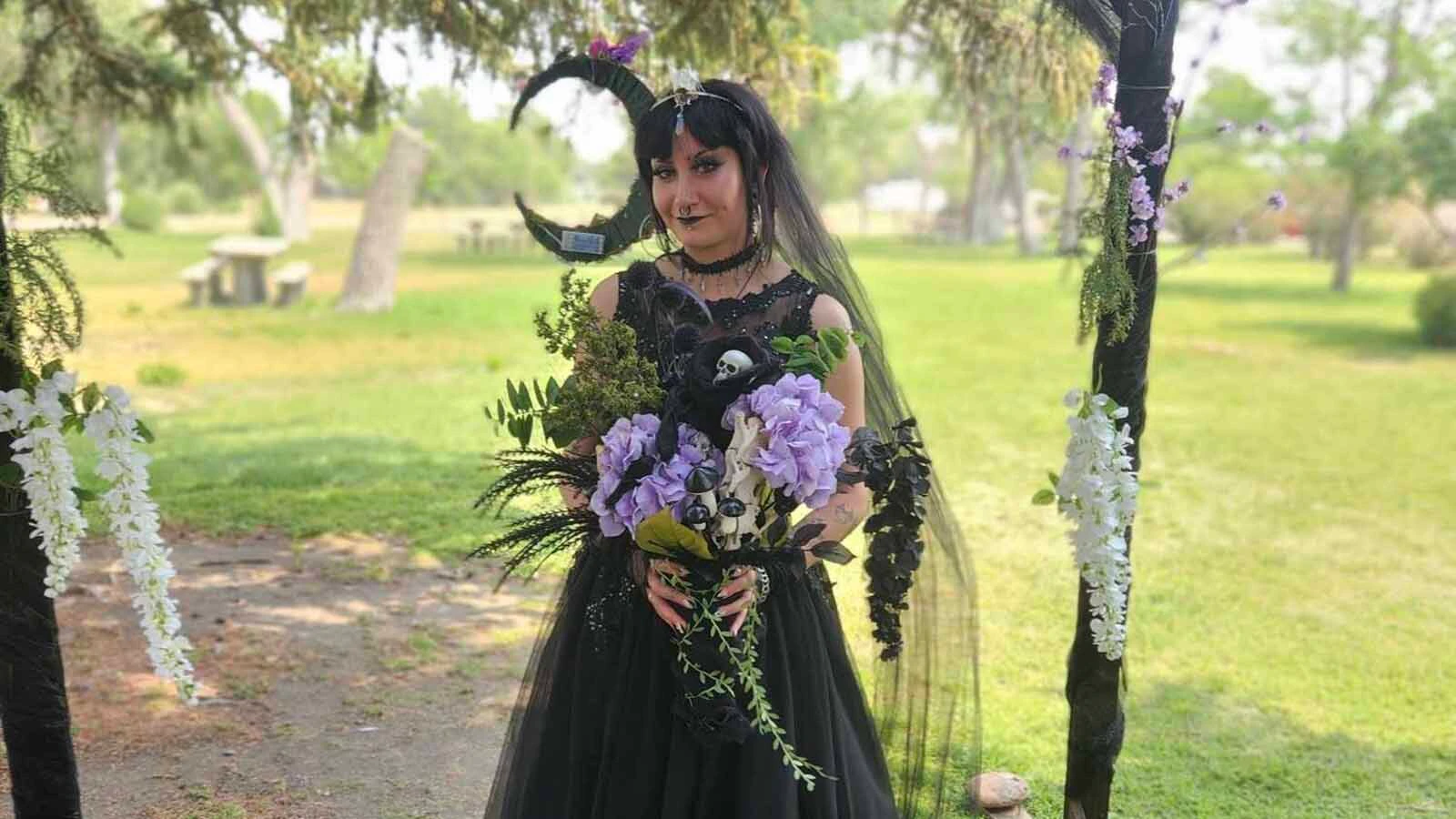 AnnChey Everhart wears a black wedding dress and veil while holding a bouquet filled with purple and black flowers, greenery and bones