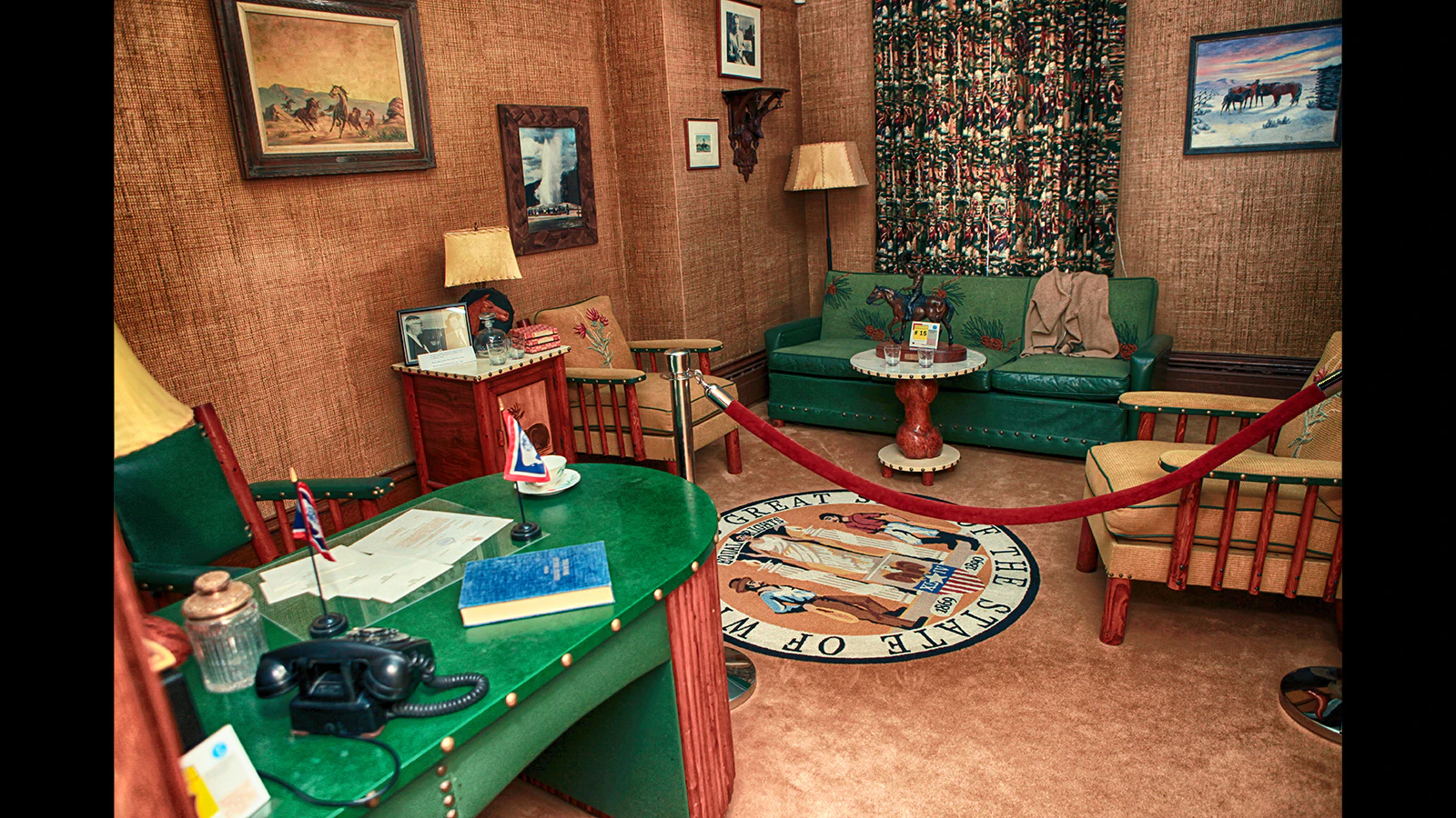 The home office in the historic Wyoming Governor's Mansion, complete with a landline phone and carpet on the walls, an emerald green and mahogany desk, some chairs, a green couch, paintings and a carpet depicting the seal of the state of Wyoming.