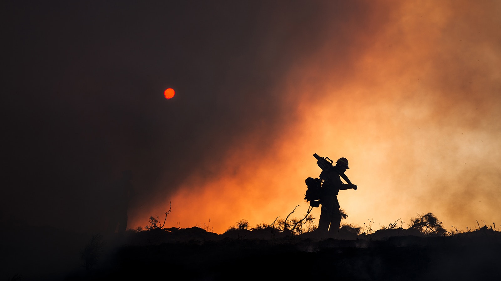 In “Sawyer Silhouette,” a wildland firefighter carries gear including a chainsaw against a backdrop of thick smoke and an orange glow. Sawyers cut down burning trees to reduce the risk of canopy fires and produce firebreaks.