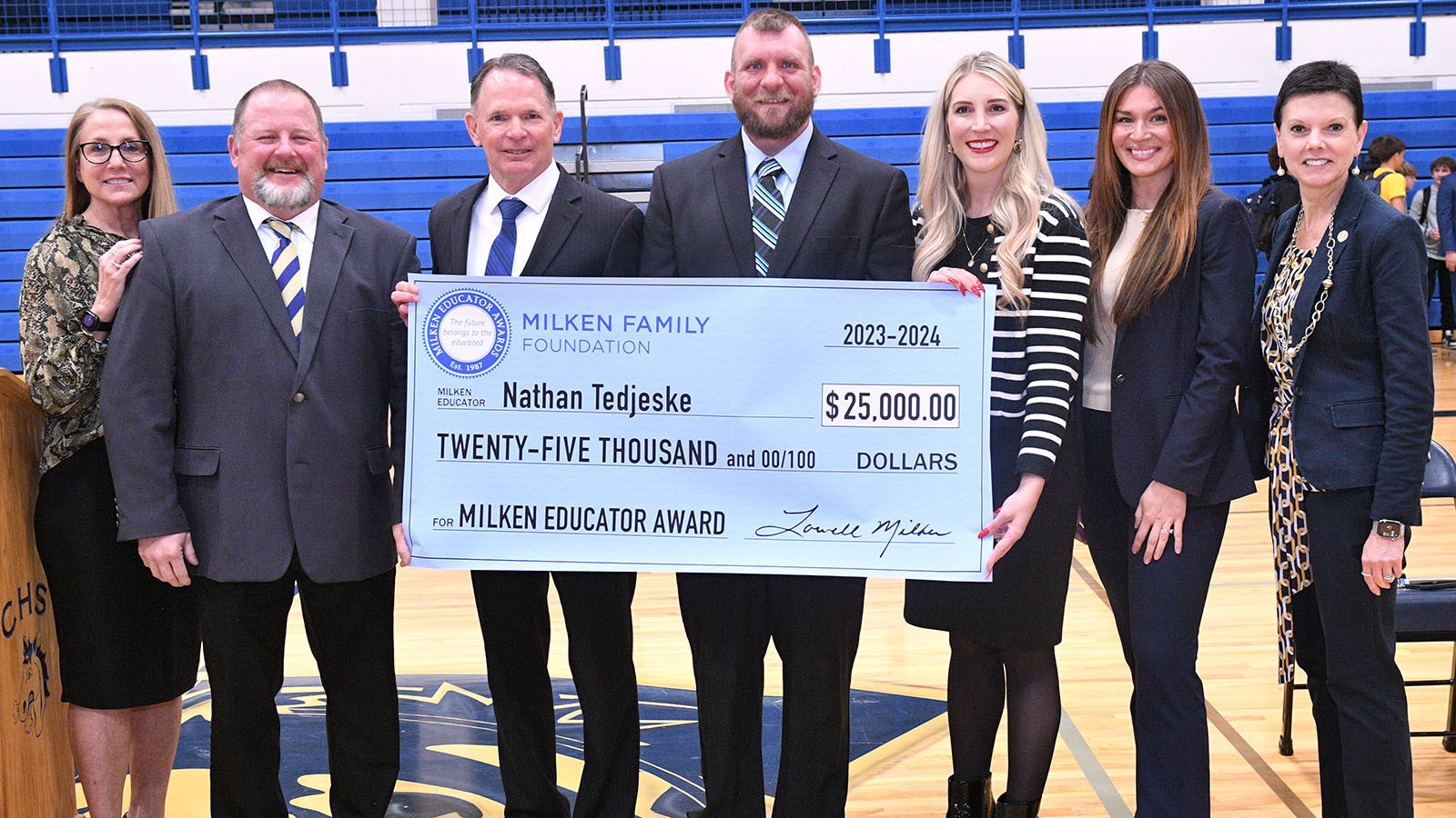 Milken Award recipient Nathan Tedjeske gets a hand with his oversized $25,000 check from visiting dignitaries and colleagues. From left, Wyoming Department of Education Communications Director Linda Finnerty; Park County School District #6 Assistant Superintendent Tim Foley; Park County School District #6 Superintendent Vernon Orndorff; recipient Nathan Tedjeske (WY '23); Wyoming Superintendent of Public Instruction Megan Degenfelder; Wyoming Department of Education Teacher Leadership and Awards Consultant Madison Lacey; and Milken Educator Awards Vice President Stephanie Bishop.