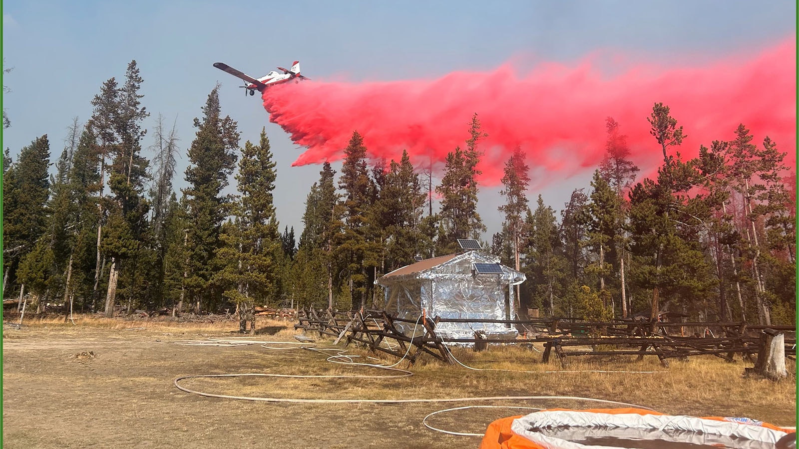 A house is wrapped in heat-resistant material as a plane drops manure above the tree line in the area of ​​the Pack Trail Fire.