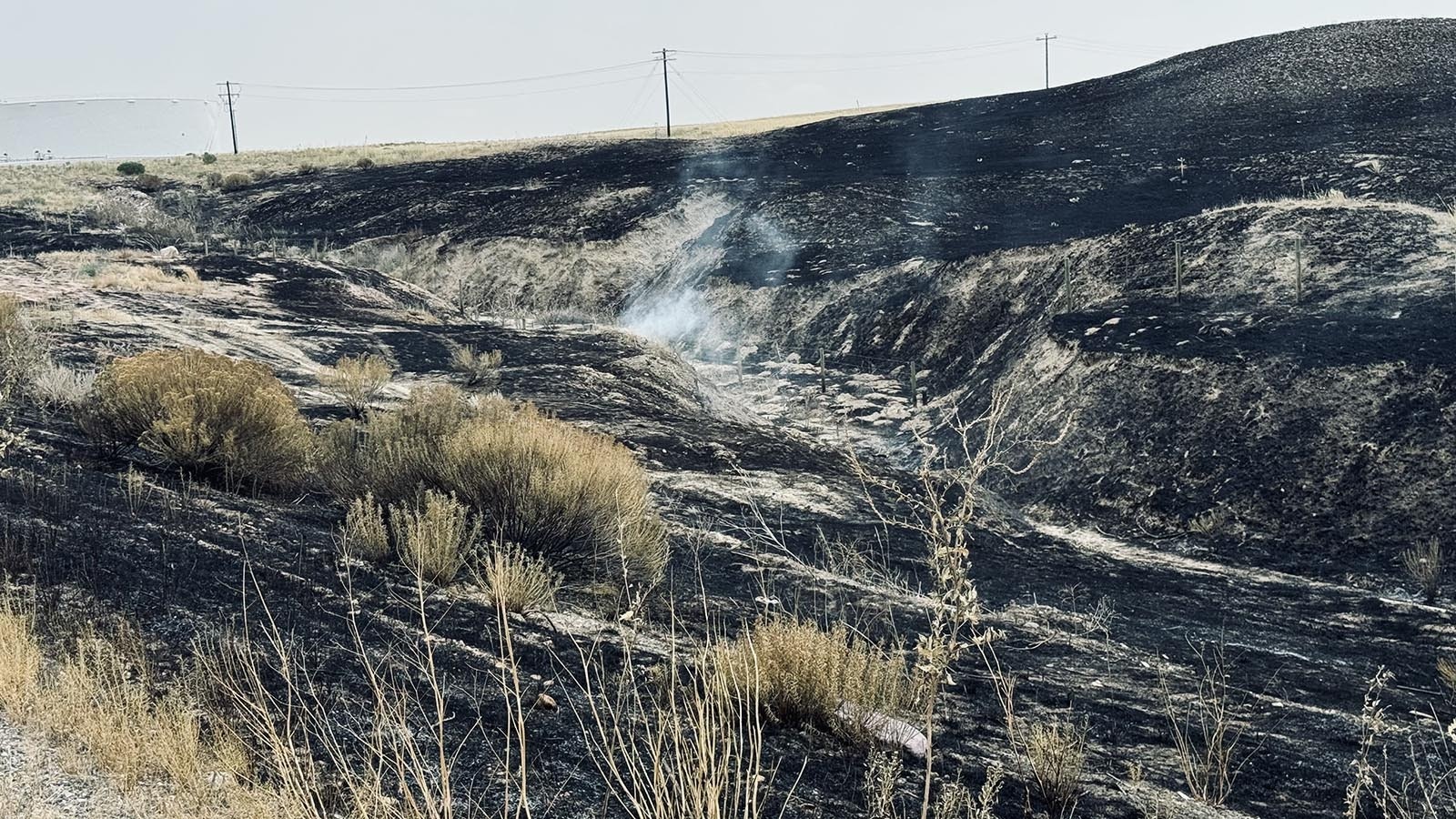 A fire smolders on Saturday south of U.S. Highway 26 near oil storage tanks owned by Tallgrass Energy in the background.