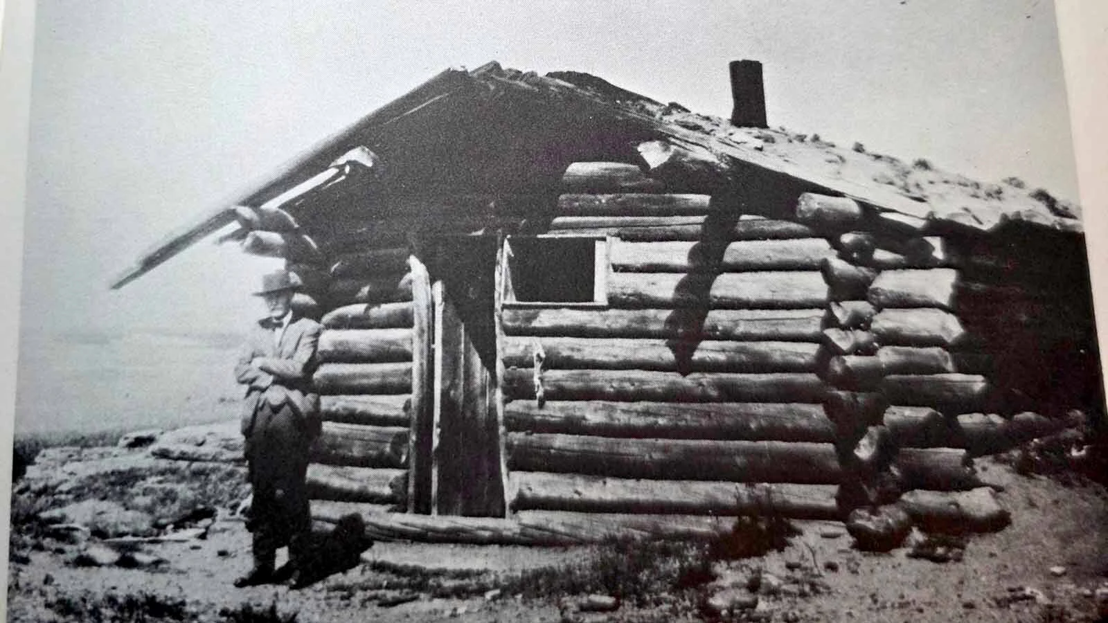 An archival photograph of prospector Bob Holland standing in front of his cabin near Bald Mountain City, the exact historic site Sara Yeaman later located by matching rock formations in the background. 