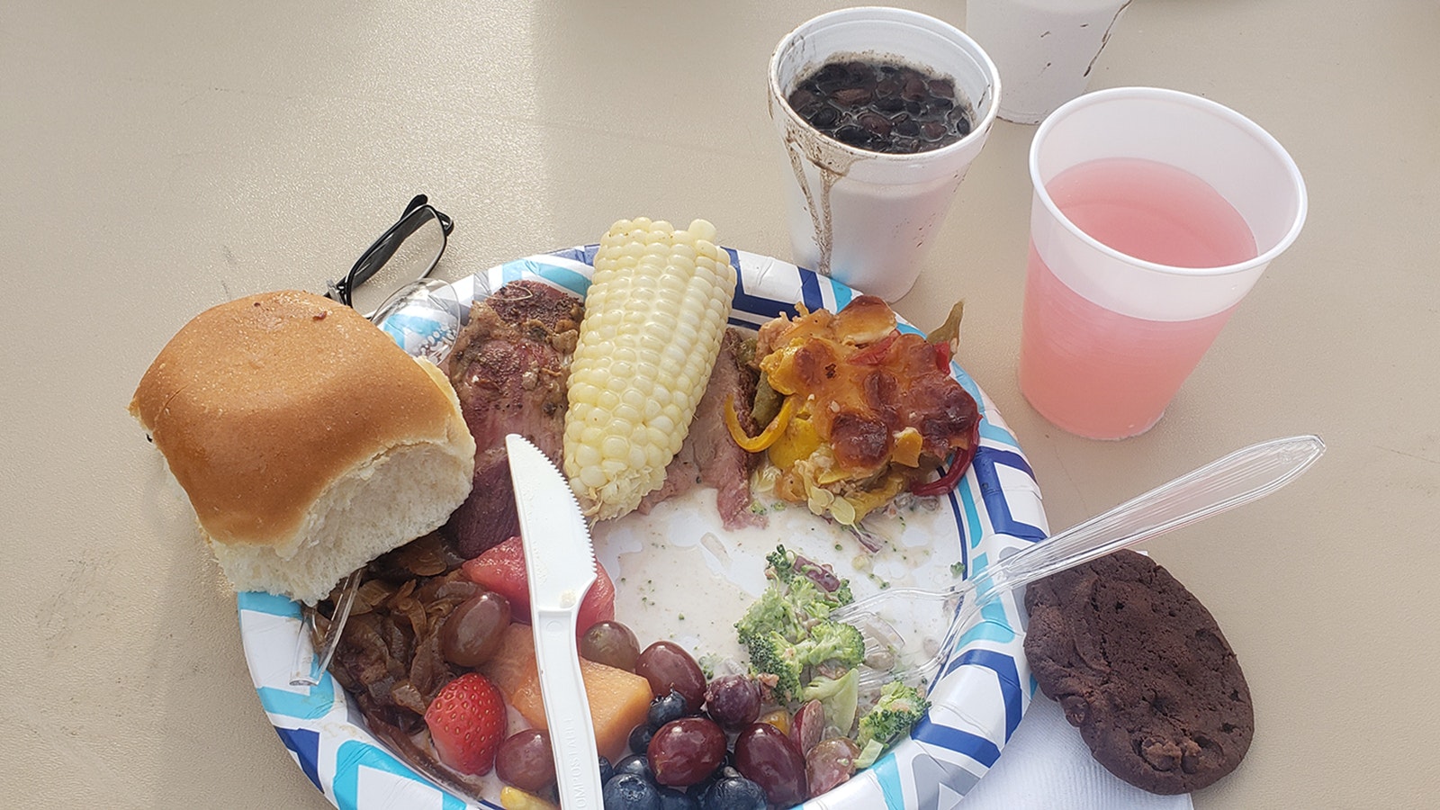 A plate with pulled pork, lamb and beef, as well as broccoli salad and squash casserole, some fruit, fresh corn and a dinner roll. The small coffee cup has beans from the famous community pot.