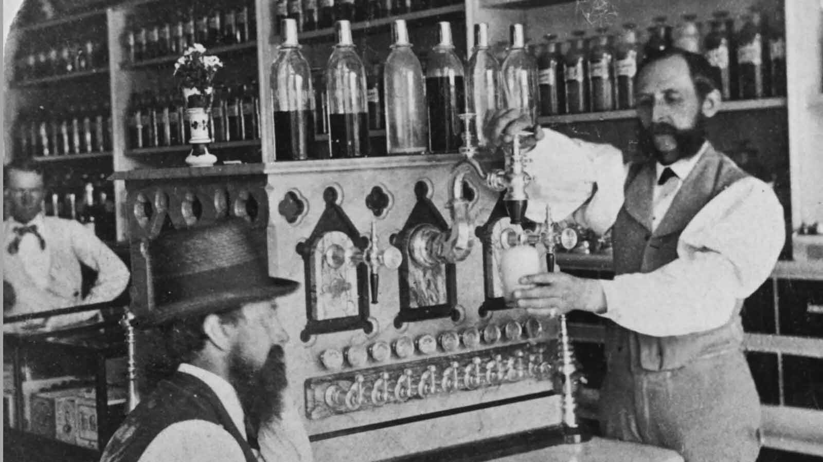 A pharmacist uses a soda fountain to prepare a drink for a customer, who sits on the other side of the counter in a drugstore, circa 1895.