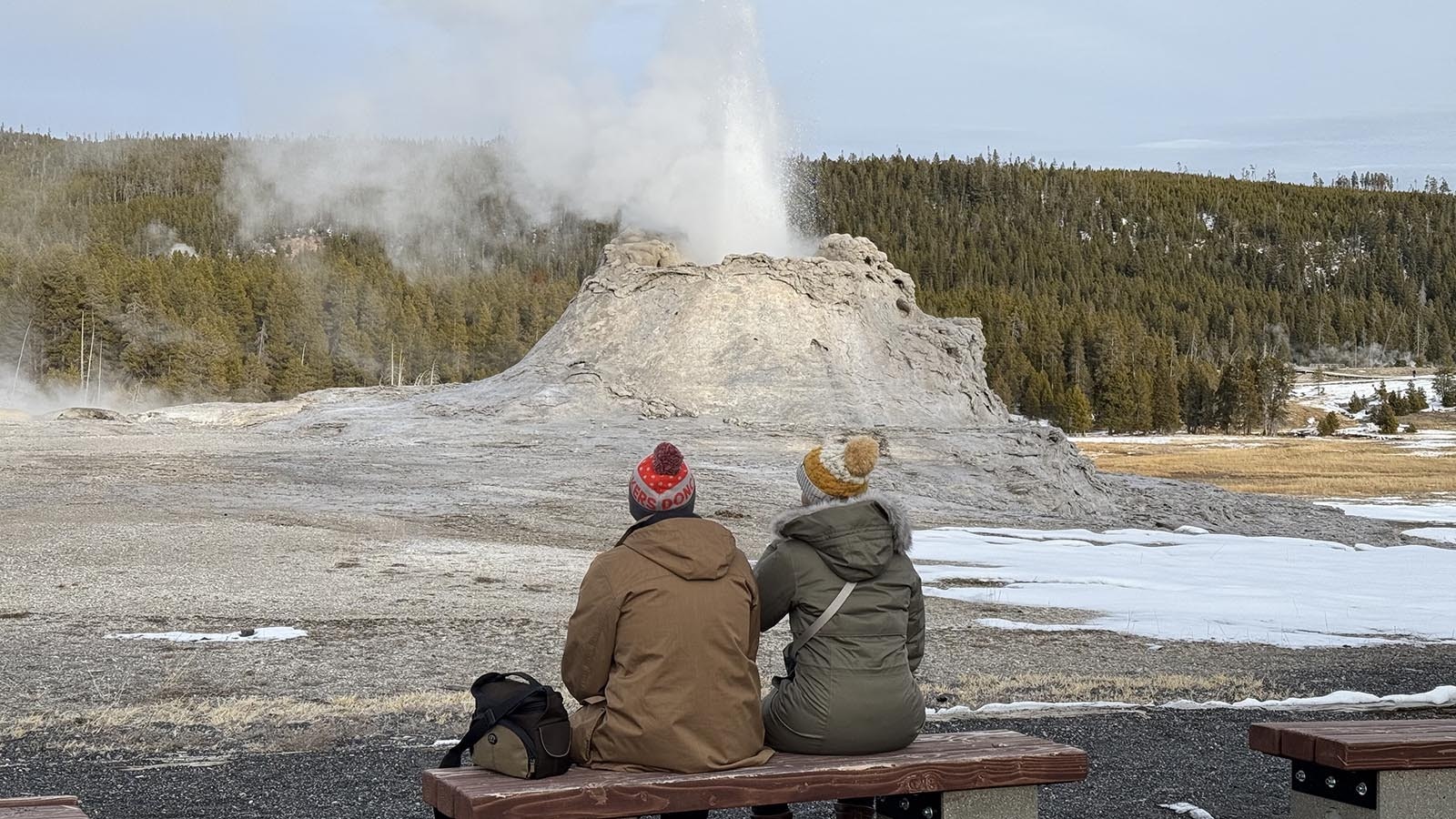 A pair of tourists in winter watch Castle Geyser in the Upper Geyser Basin of Yellowstone National Park.