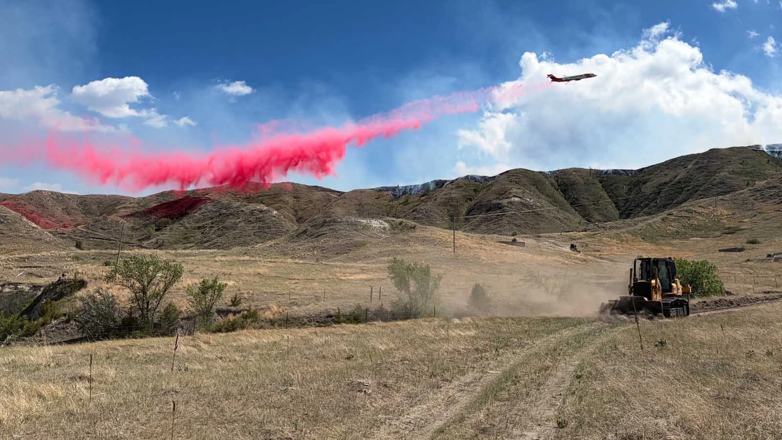 Pink fire retardant streams from an air tanker above a dozer carving a containment line during a wildfire operation.
