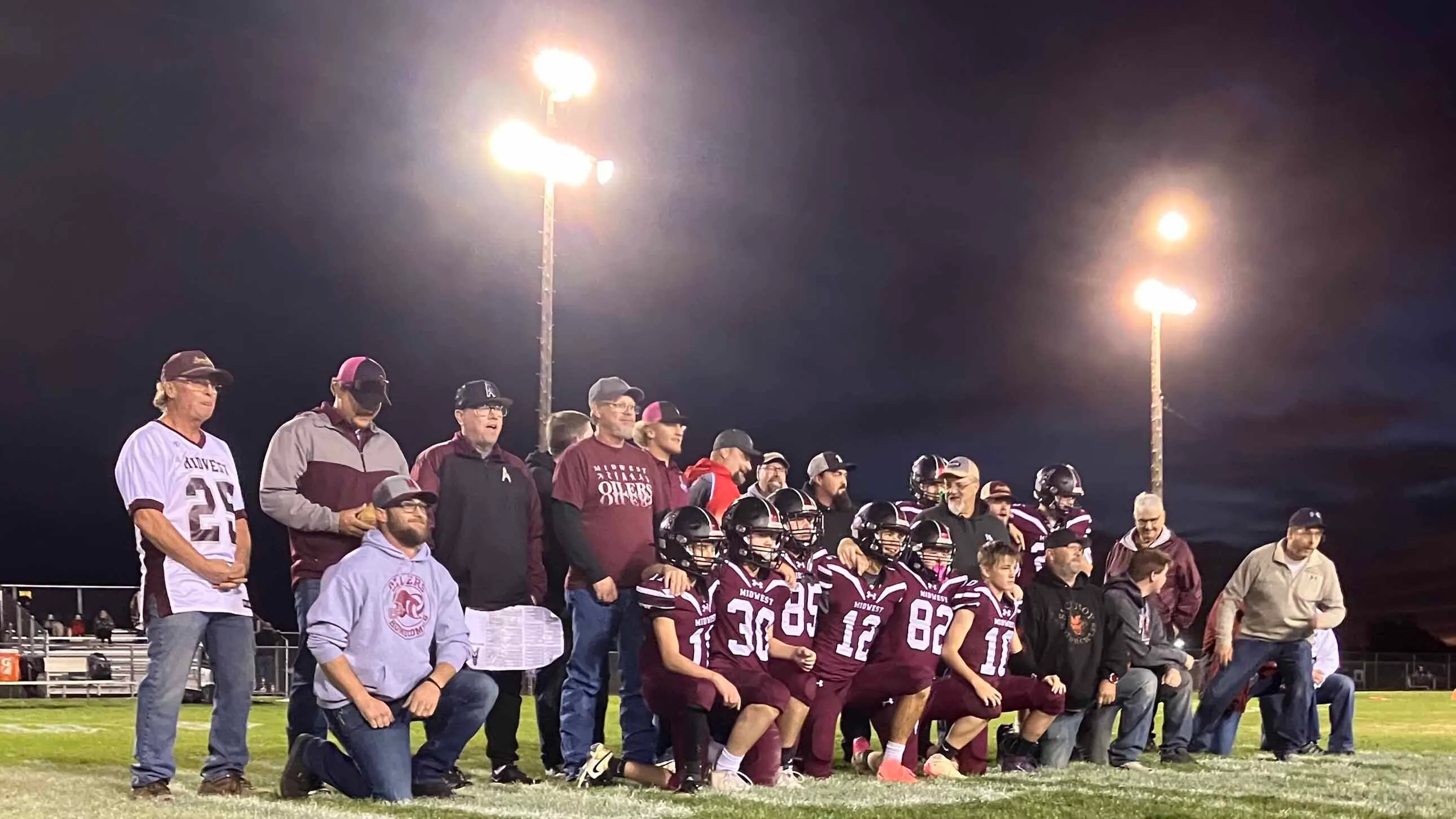Members of the 2025 Midwest Oilers football team, along with squad alumni, pose for a photo together at halftime of a game commemorating the 100th anniversary of playing night football under lights.