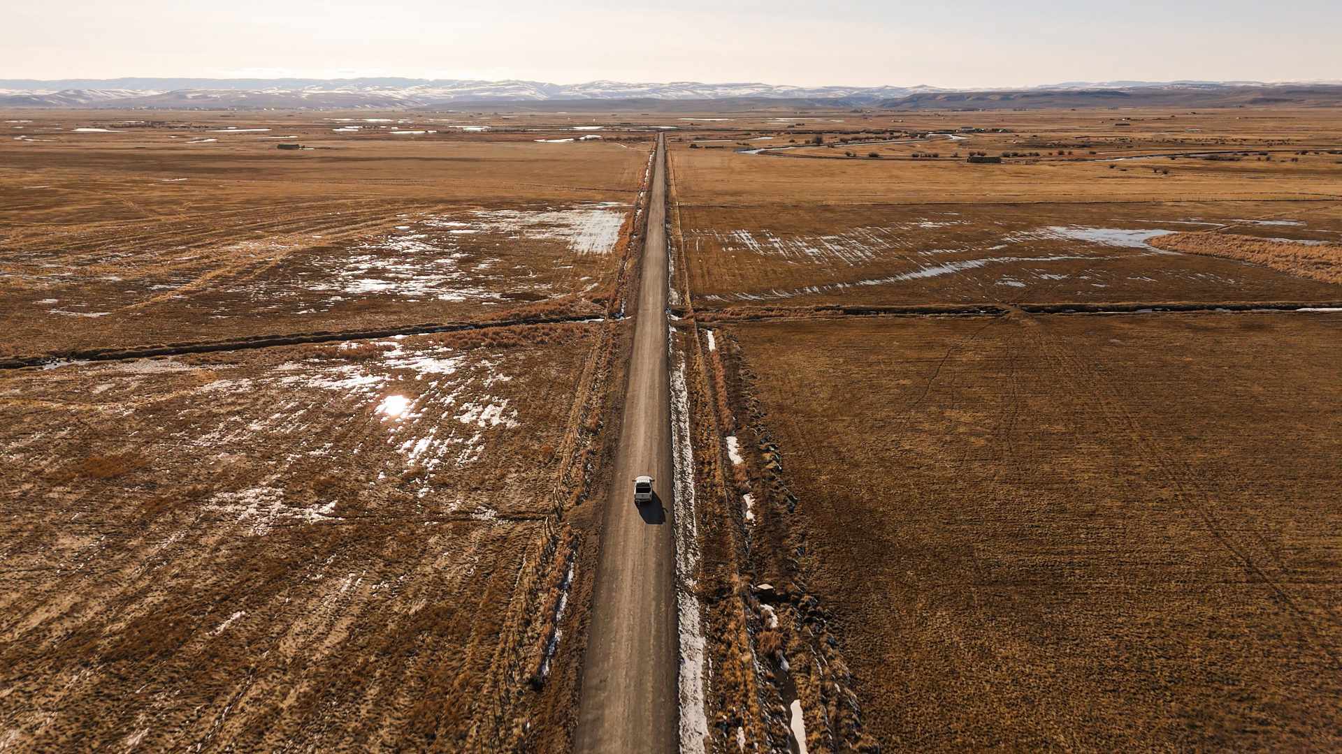 A vehicle is driving into the horizon along a straight dirt road from an overhead view.